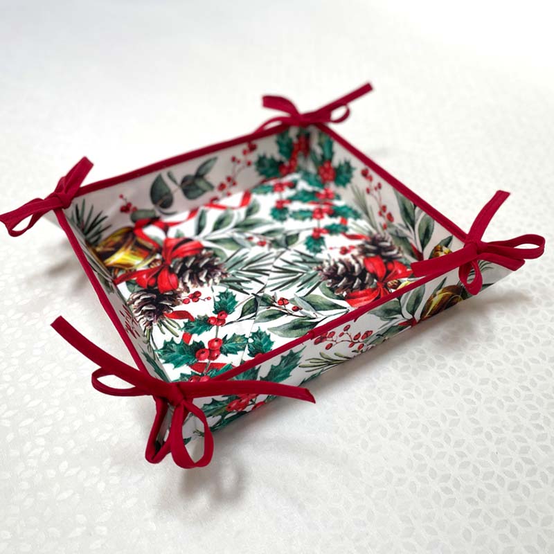 A square bread basket with a white background and a festive holly and pine print, finished with red ribbon ties at the corners, photographed on a white tablecloth.