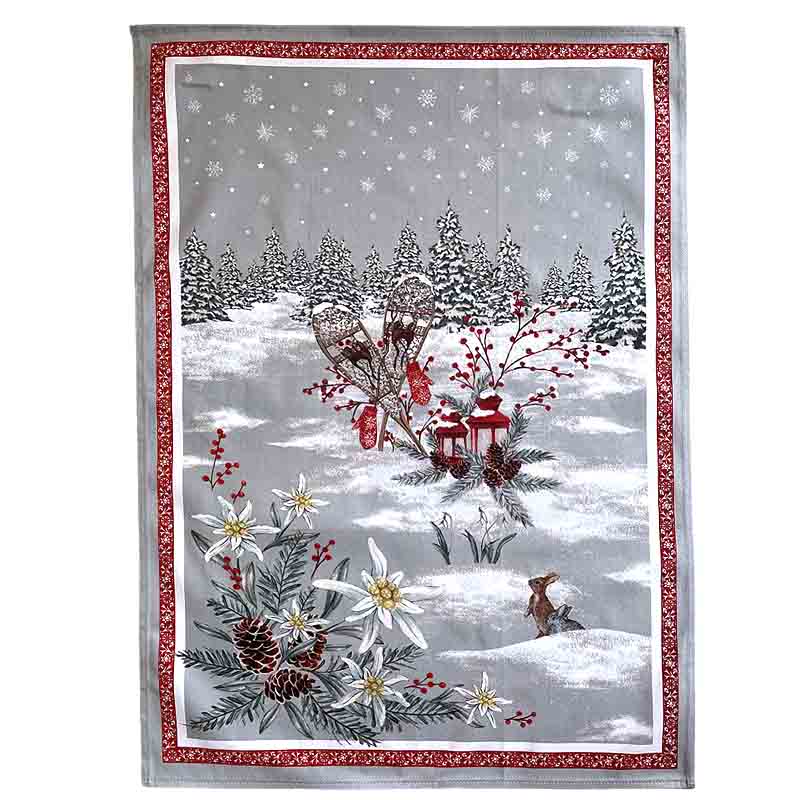A rectangular holiday-themed tea towel featuring a snowy winter landscape with evergreen trees, white flowers, red berries, lanterns, and a small deer, framed by a red patterned border on a light gray background.