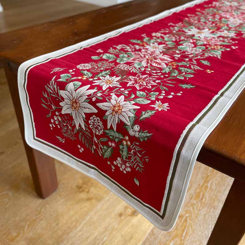 A red table runner with a festive floral pattern of white poinsettias, green leaves, and small birds, draped over a dark wooden table on a light wood floor.