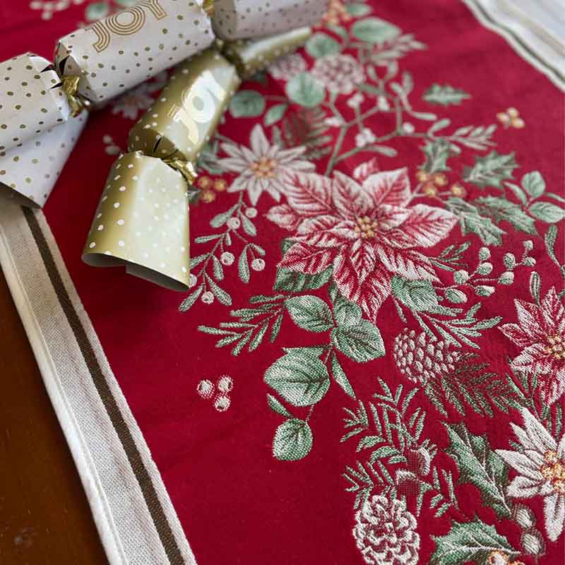 A close-up of a red table runner with a festive embroidered floral pattern in pink, white, and green, styled with gold and white polka dot cracker fireworks on a wooden surface.
