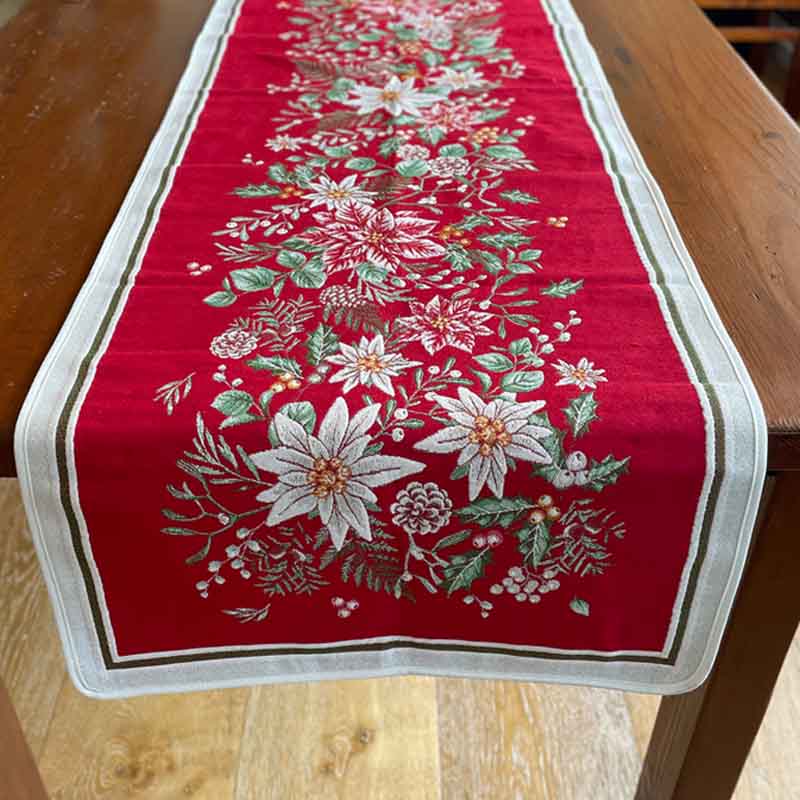 A festive Christmas table runner with a vibrant red background, featuring a detailed floral and botanical pattern in white, green, and gold, finished with a white border.