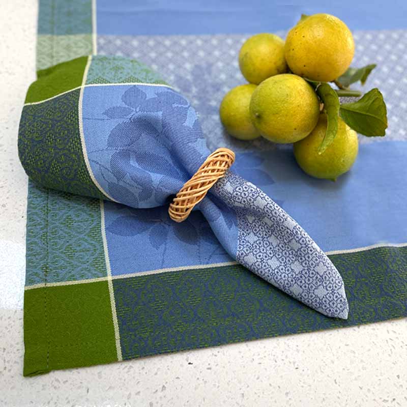 Blue and green patterned napkin and tablecloth with lemons and a woven napkin ring on a white surface.