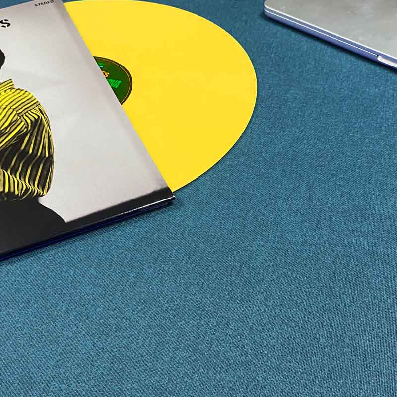 A close-up of a textured blue fabric tablecloth with a yellow vinyl record partially tucked under a record sleeve, alongside part of a silver laptop.