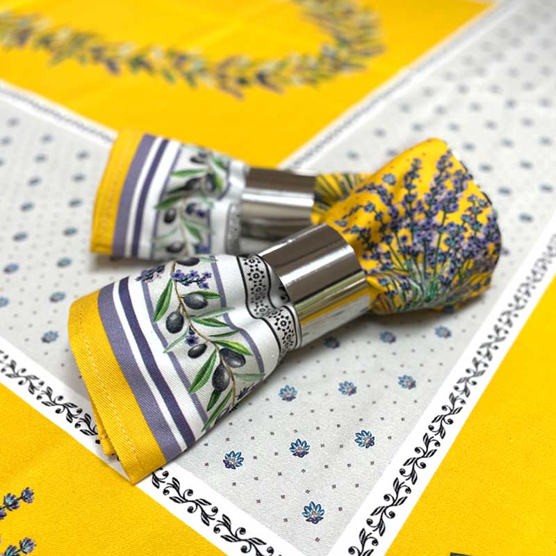 Two metal napkin rings holding patterned napkins, placed on a yellow and white patterned tablecloth. The napkin designs feature olive branches and lavender sprigs.