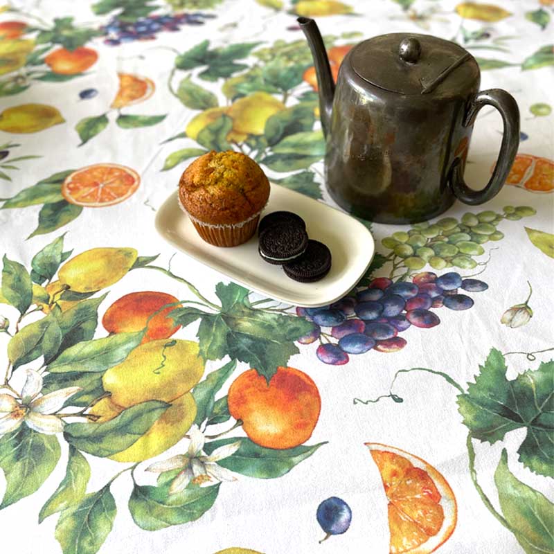 Bright summer fruits design on a tablecloth with a tea pot and a muffin and biscuits on a plate.
