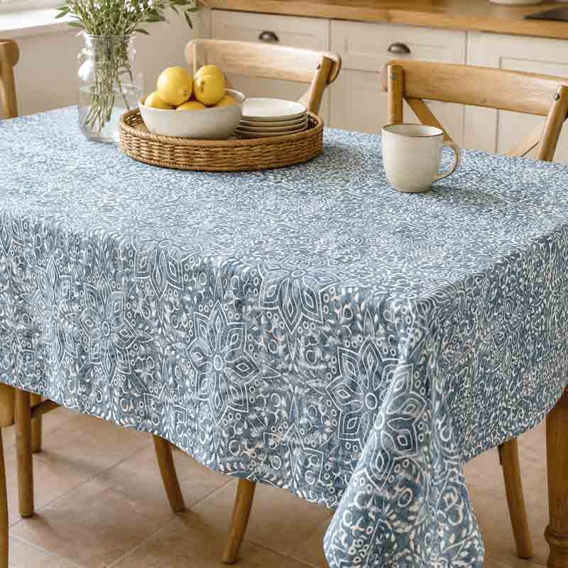 A rectangular table covered with a light blue and white patterned tablecloth, set in a bright kitchen with wooden chairs, a wicker tray with lemons, stacked plates, and a ceramic mug.