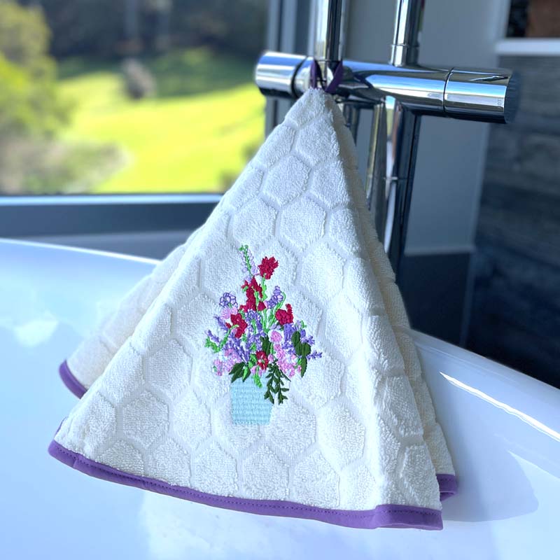 A cream textured hand towel with a purple border and a colorful embroidered flower bouquet design, hanging from a chrome faucet on a white bathtub with a blurred outdoor background.