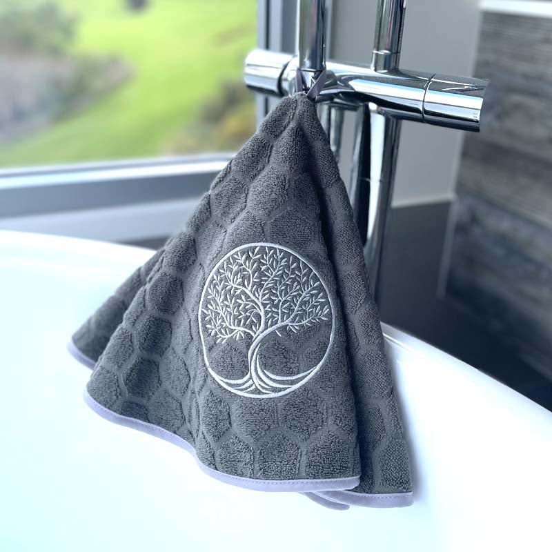 A gray hand towel with a silver embroidered 'Tree of Life' design, hanging from a chrome faucet in a bathroom setting.