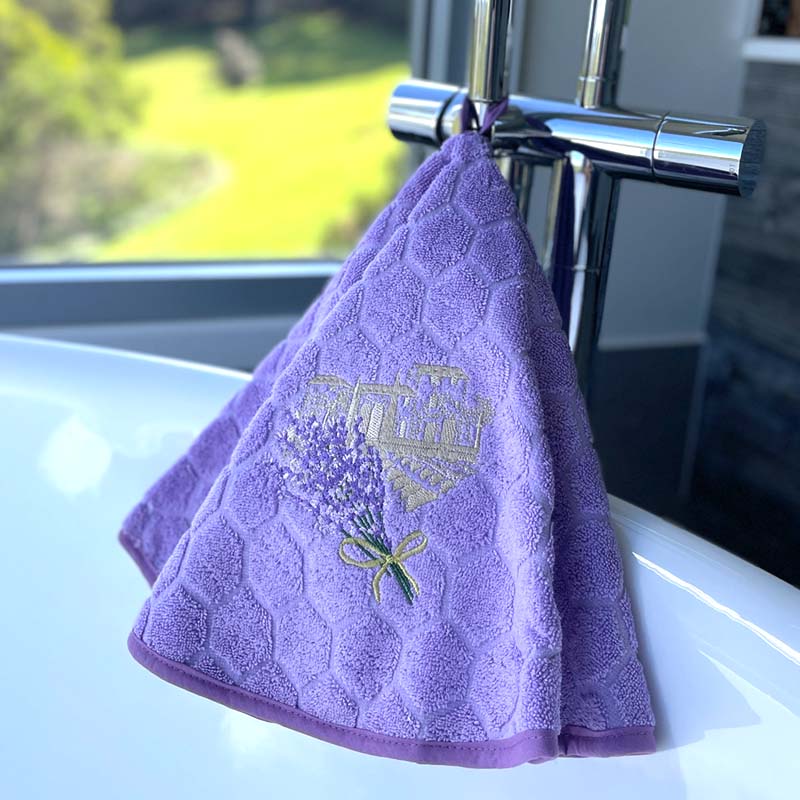 A purple textured hand towel with an embroidered lavender bouquet and gold bastide design, hanging from a chrome faucet on a white bathtub with a blurred outdoor background.