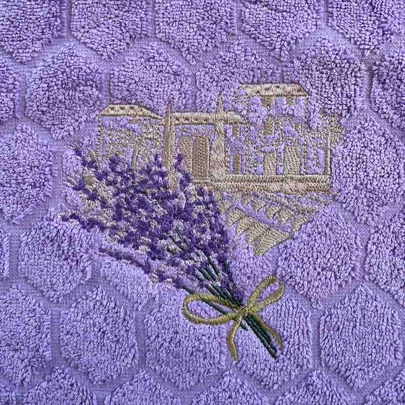 A close-up of a lavender-themed hand towel featuring a textured purple background with a raised floral pattern, embroidered lavender sprigs, and a gold-toned bastide motif.