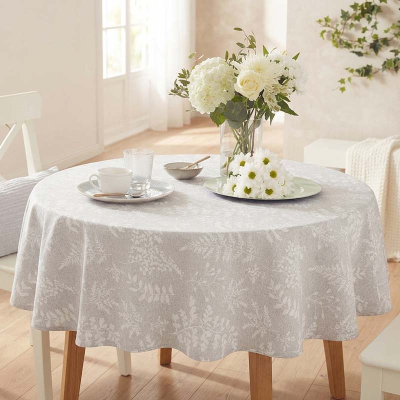 A round dining table covered with a light gray and white botanical patterned tablecloth, styled with a vase of white flowers, a teacup, and a glass in a bright, softly lit room.