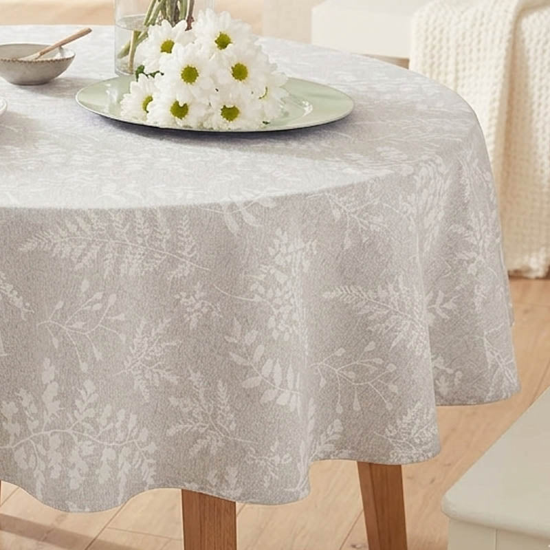A round table covered with a light gray tablecloth featuring a subtle white botanical leaf pattern. A plate of white daisies and a small bowl are placed on the table, with a softly blurred indoor room background showing light wood flooring and a cream chair.