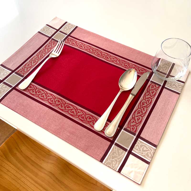 Red placemat with decorative border on a wooden table, featuring silver cutlery and a glass.