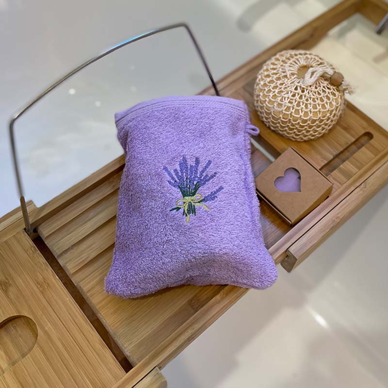 A lavender-colored textured bath mitt with an embroidered lavender bouquet on the front, resting on a wooden tray next to a woven pouch and a small wooden box.
