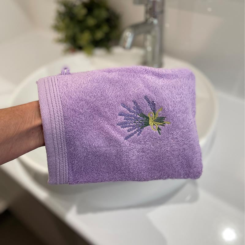 A lavender purple bath mitt with a small embroidered lavender and green flower motif, held by a hand in front of a white ceramic sink.