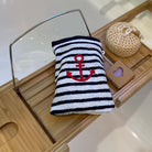 A striped bath mitt with a red embroidered anchor, displayed on a wooden tray alongside a woven bag and a small wooden heart box.