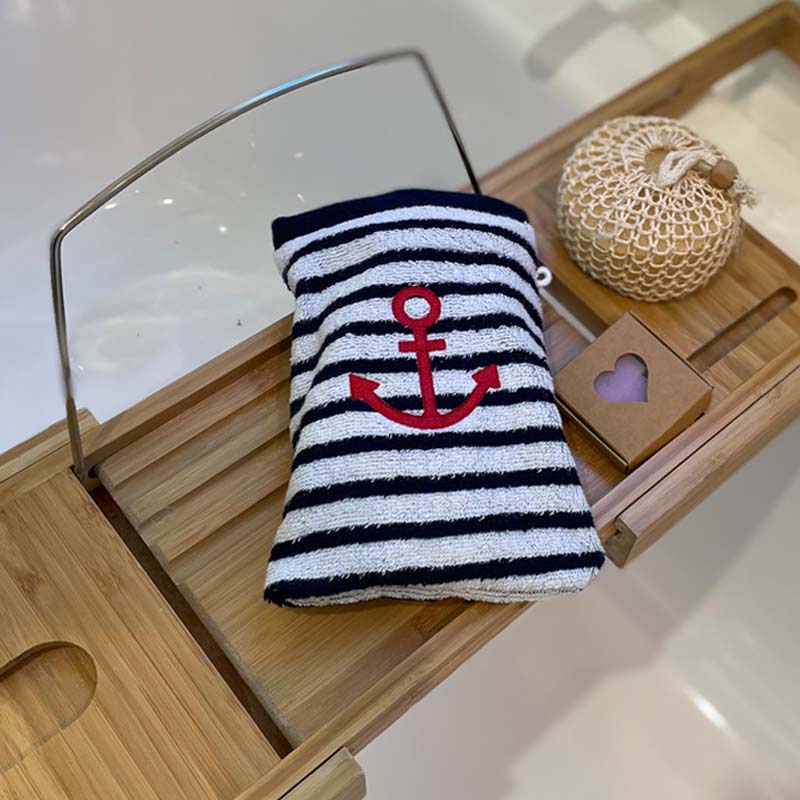 A striped bath mitt with a red embroidered anchor, displayed on a wooden tray alongside a woven bag and a small wooden heart box.