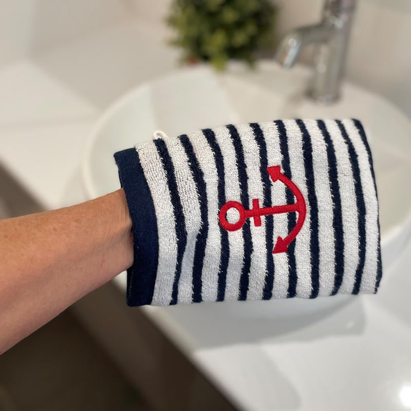 A close-up of a person's arm wearing a white terry cloth bath mitt with navy blue vertical stripes and a red embroidered anchor, held over a white sink in a bathroom setting.