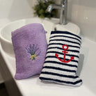 Two bath mitts on a white sink: one lavender purple bath mitt with an embroidered lavender bouquet, and one navy and white striped with a red anchor.