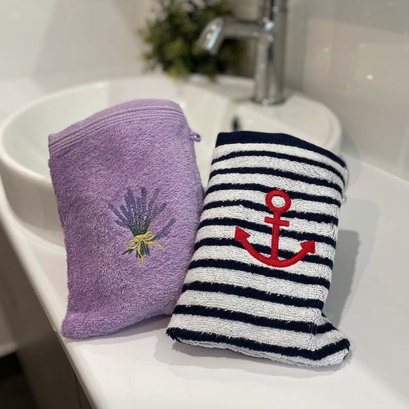 Two bath mitts on a white sink: one lavender purple bath mitt with an embroidered lavender bouquet, and one navy and white striped with a red anchor.