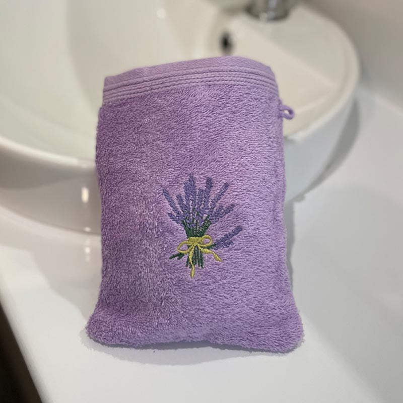 A lavender purple cotton washcloth pouch with an embroidered lavender bouquet on the front, resting on a white bathroom sink counter.