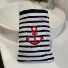A washcloth bath mitt with navy and white horizontal stripes and a red anchor motif, placed on a white sink countertop.