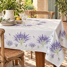 A light grey background tablecloth with a repeating purple lavender bouquet pattern, draped over a wooden dining table set with plates, glasses, and a pitcher in a sunlit outdoor patio.