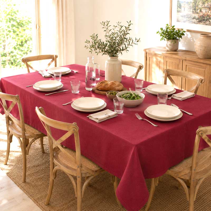 A rectangular dining table covered with a deep red tablecloth, set with white plates, glasses, cutlery, a loaf of bread, a bowl of salad, and a vase of greenery. The table is surrounded by light wood chairs in a bright, sunlit dining room.