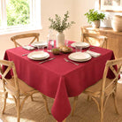 A red tablecloth draped over a rectangular dining table set with white plates, silverware, glasses, and a vase of greenery, surrounded by wooden chairs in a bright dining room.