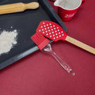 A pastry brush and a spatula arranged on the deep red coloured Bordeaux tablecloth next to a baking tray, flour, a wooden rolling pin, and a red measuring cup.