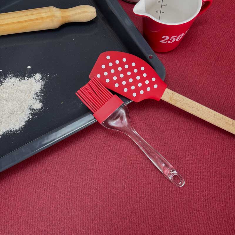 A pastry brush and a spatula arranged on the deep red coloured Bordeaux tablecloth next to a baking tray, flour, a wooden rolling pin, and a red measuring cup.