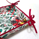 A square bread basket with a white background and a festive Christmas botanical print featuring green leaves, red berries, pinecones, and gold bells, finished with a red ribbon border and tied bows.