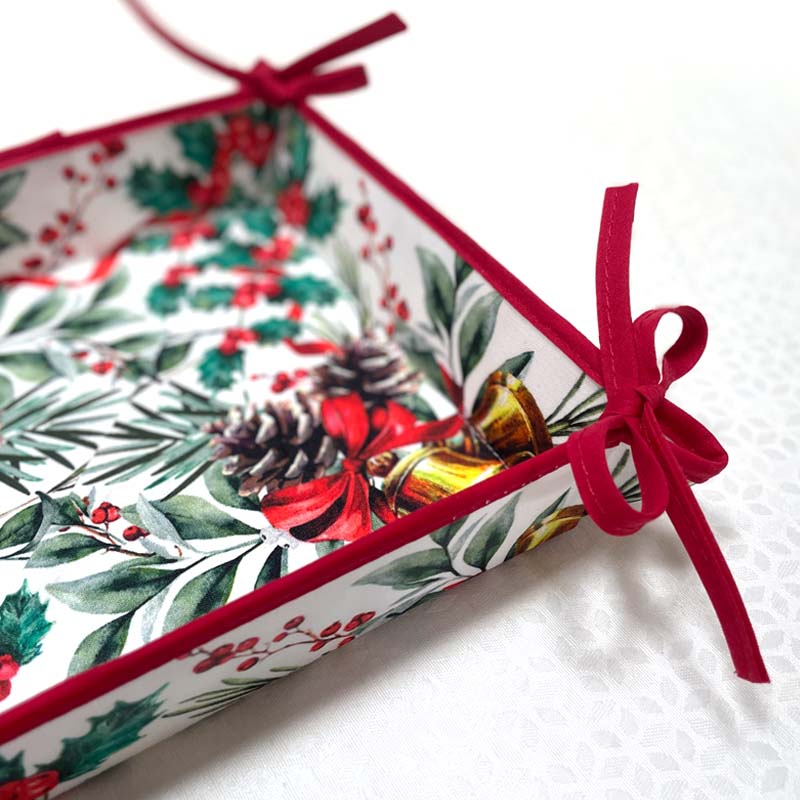 A square bread basket with a white background and a festive Christmas botanical print featuring green leaves, red berries, pinecones, and gold bells, finished with a red ribbon border and tied bows.