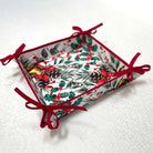 A square bread basket with a white background and a festive holly and pine print, finished with red ribbon ties at the corners, photographed on a white tablecloth.