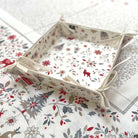 A square fabric storage basket with a festive Christmas print, featuring red reindeer, holly, and snowflakes on an off-white background, styled on top of a matching patterned textile.
