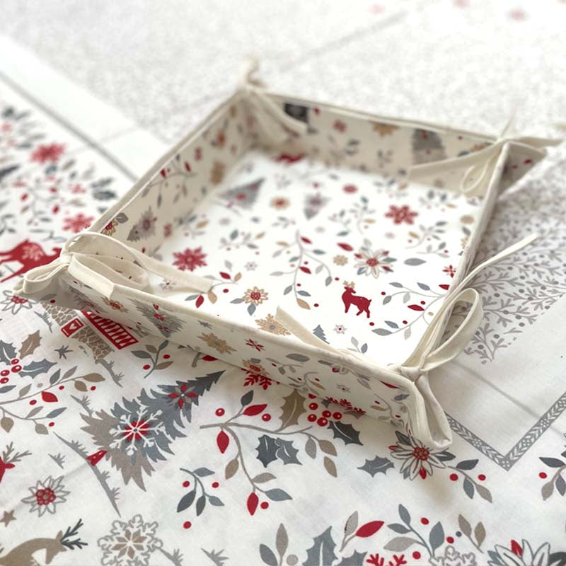 A square fabric storage basket with a festive Christmas print, featuring red reindeer, holly, and snowflakes on an off-white background, styled on top of a matching patterned textile.