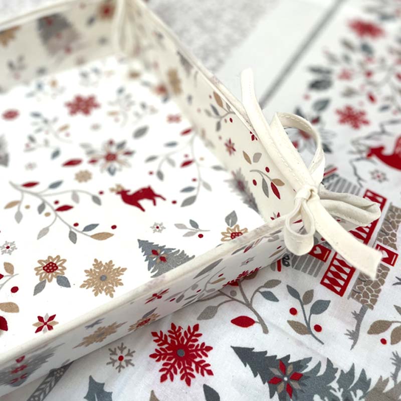 A close-up of a white bread basket in festive Christmas patterned fabric with red, gray, and beige holiday motifs, tied with a cream ribbon.