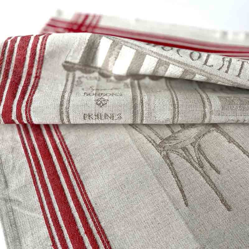 A folded linen kitchen towel with red and white stripes and a gray printed design, showing the brand name 'Dormont & Company' and 'Cannes' on the fabric.
