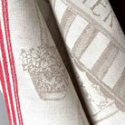 Close-up of a woven jacquard tea towel, in off-white with a taupe basketweave pattern, featuring a floral motif and red and white stripes along one edge.