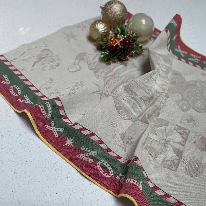 A Christmas tea towel with a festive border and subtle holiday motifs, styled on a white speckled countertop with decorative ornaments.