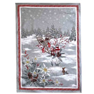 A rectangular holiday-themed tea towel featuring a snowy winter landscape with evergreen trees, white flowers, red berries, lanterns, and a small deer, framed by a red patterned border on a light gray background.