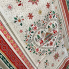 A close-up of a tea towel featuring a festive Christmas print with a central heart-shaped holiday scene, surrounded by floral motifs and bordered by red, green, and gold patterned trim.