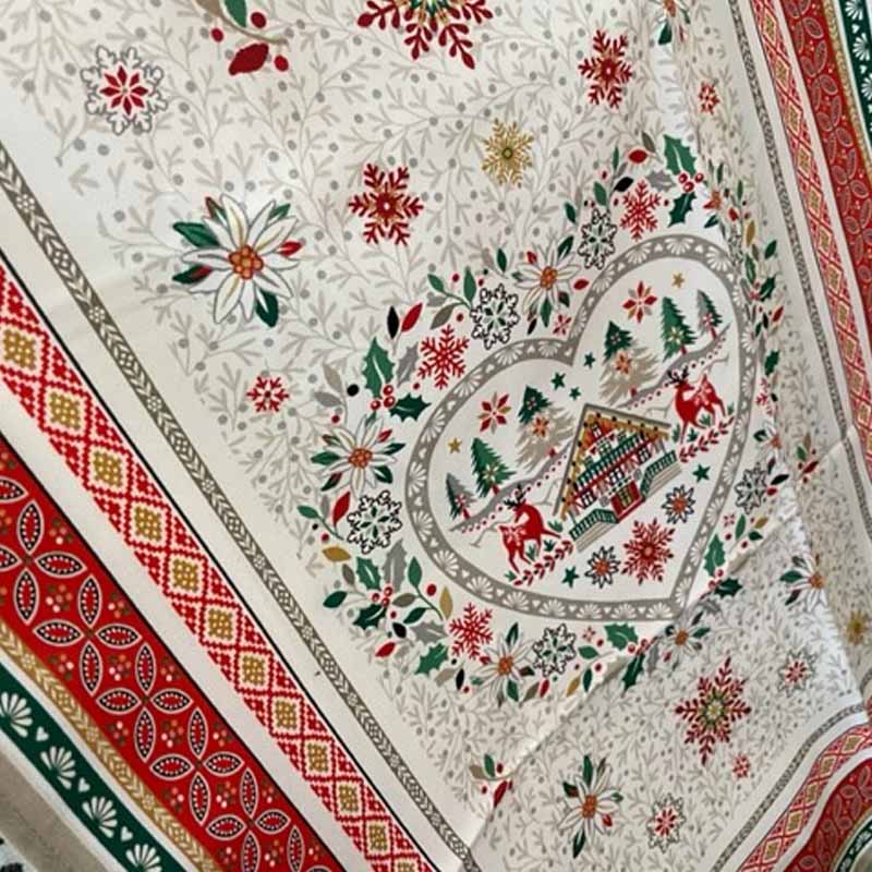 A close-up of a tea towel featuring a festive Christmas print with a central heart-shaped holiday scene, surrounded by floral motifs and bordered by red, green, and gold patterned trim.