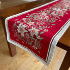 A red table runner with a festive floral pattern of white poinsettias, green leaves, and small birds, draped over a dark wooden table on a light wood floor.