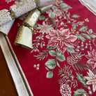 A close-up of a red table runner with a festive embroidered floral pattern in pink, white, and green, styled with gold and white polka dot cracker fireworks on a wooden surface.
