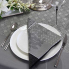 Dining table setting with a patterned napkin, white plates, silverware, and flowers on a gray patterned tablecloth.