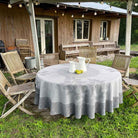 Round table with a patterned tablecloth in an outdoor setting with wooden chairs and a cabin in the background.