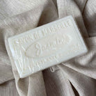 A rectangular bar of Savon de Marseille Jasmine soap wrapped in clear plastic, resting on a textured beige linen fabric background.