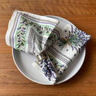 Two folded patterned napkins on a white plate over a wooden table. One napkin has green olive branches with black and white detailing, and the other has purple lavender sprigs with green stems and a small twine bow.