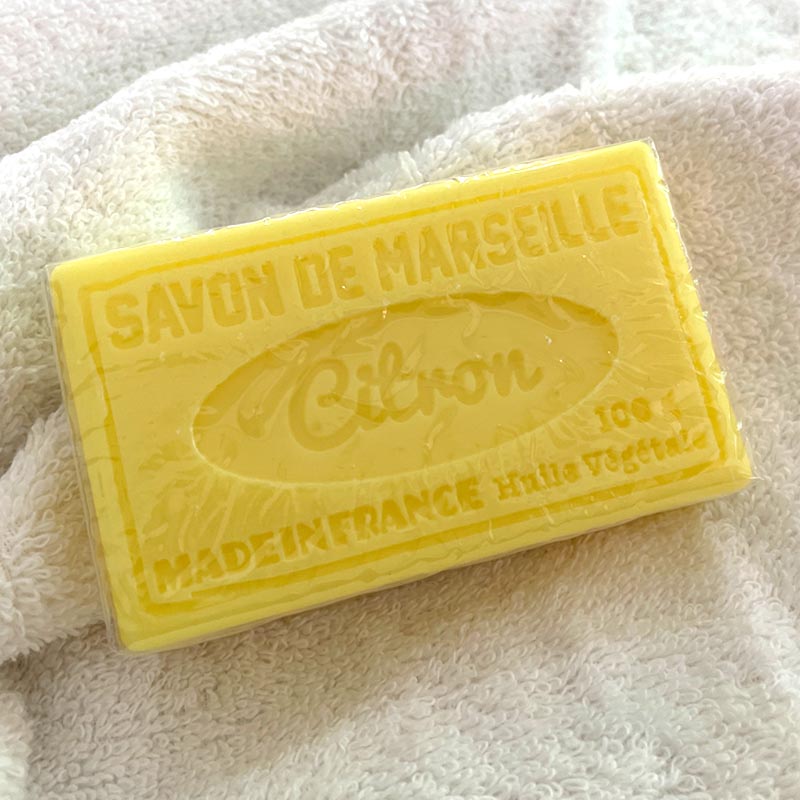 A rectangular bar of yellow citron soap resting on a soft white textured towel. The soap features embossed text reading 'SAVON DE MARSEILLE Citron' and 'MADE IN FRANCE'.